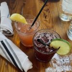Two colorful non-alcoholic mocktails served on a wooden table at Tazza Kitchen in Richmond, Virginia. The drinks, "True Story" and "Sunday Best," are in short glasses with ice and garnishes—one with a lemon wedge and the other with a lime slice. Black straws are in both glasses, and white cloth napkins with black stripes are folded nearby. A glass of ice water is visible in the background.