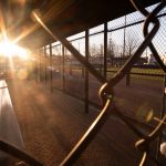 The sun reflects off a metal bench in a dugout, surrounded by chain link fence.