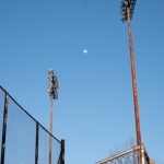 Two stadium lights rise above the backstop, with the moon between them on a clear blue sky.
