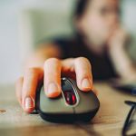 A hand is resting on a computer mouse on a wooden table, with cords on the right side.