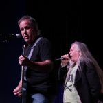 A man to the left is pictured singing while playing the guitar as a man to the right sings in a stage.