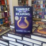 A hardcover copy of Sunrise on the Reaping by Suzanne Collins sits on a table at Barnes & Noble in Knoxville, TN, on its release day. The book's cover features a golden mockingjay against a dark background. Other copies are stacked nearby, with store shelves visible in the background.