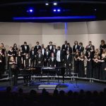 A full choir ensemble of young men and women stand on risers singing.