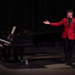 A man in a red suit gestures to his accompanist at the end of his performance, queuing a bow.