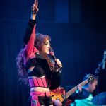Chapple Roan singing at The Vogue Theatre, arm raised in the air holding up two fingers