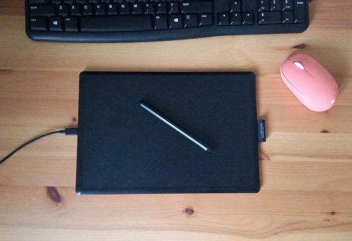 A drawing tablet, a typing keyboard, and a computer mouse lay on a wooden table.