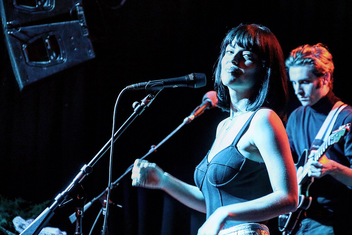 Woman stands at a microphone stand with guitar player in the background.
