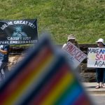 Pride flag objects view of protestors across the street holding signs reading "make America sane again" and "are we great yet? 'cause I just feel embarrassed."
