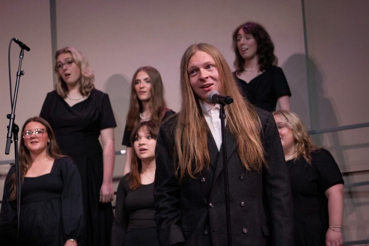 Will Dishmon in a tuxedo passionately sings in front of a microphone with the choir behind him.