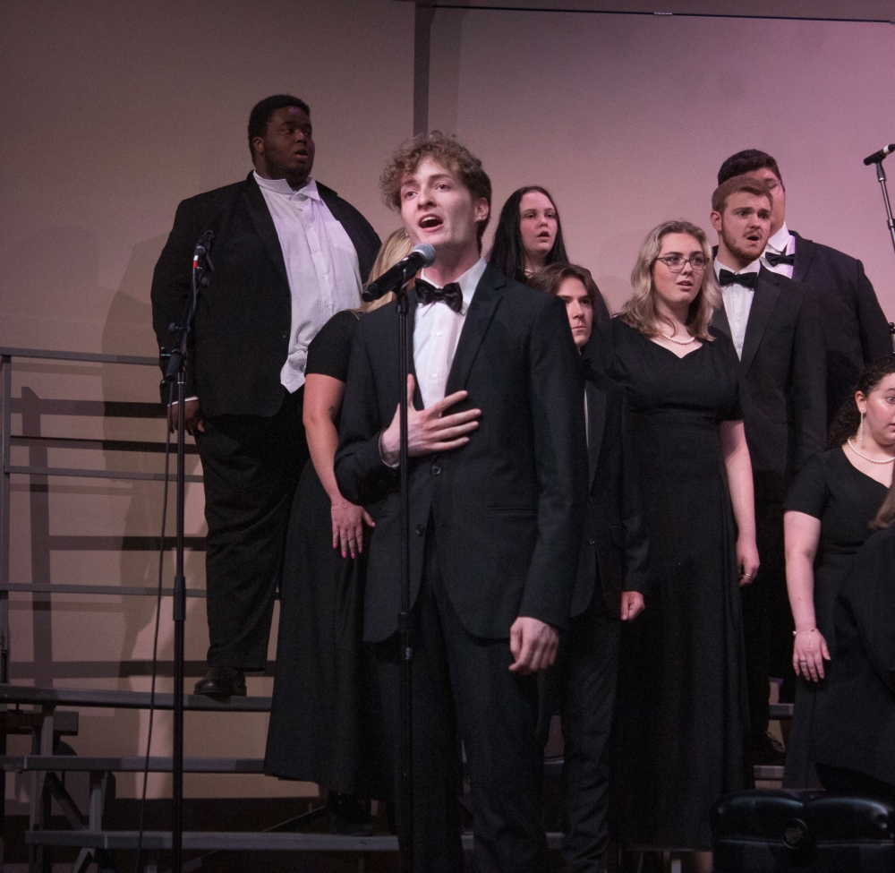 Isaiah Crisp in a tuxedo passionately sings in front of a microphone with the choir behind him.