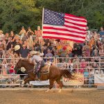 A woman is riding a horse, holding an American Flag and riding through the dirt stadium