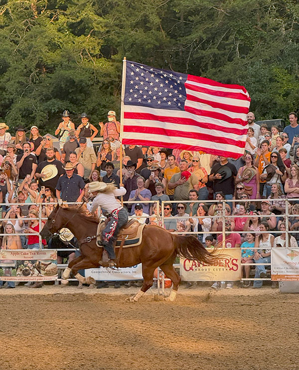 A woman is riding a horse, holding an American Flag and riding through the dirt stadium