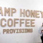 A woman stands next to a sign that says "Camp Honey Coffee"