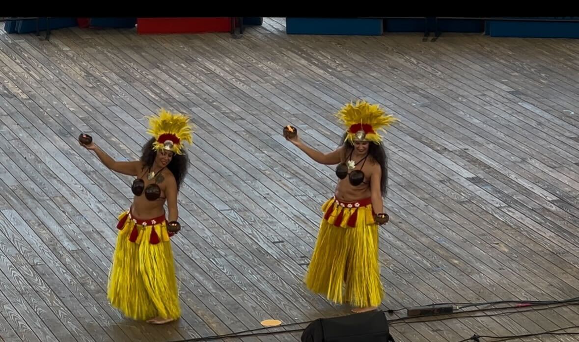 Two female Polynesian dancers in traditional yellow garb dance on a stage.