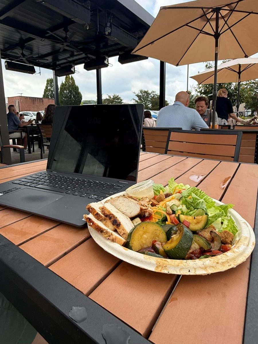 A laptop next to a plate full of chicken, veggies, and a salad at an outdoor seating area