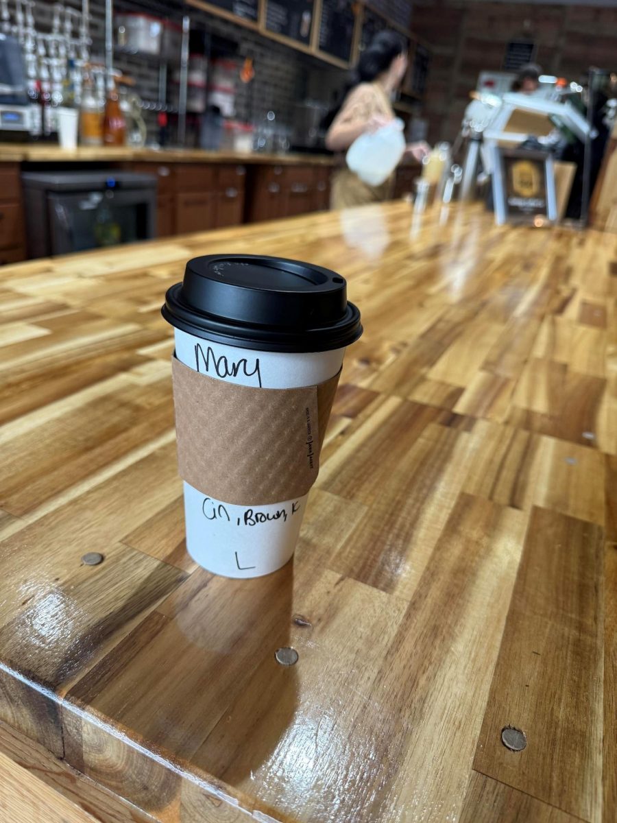 a cup of coffee on a wooden counter with Mary's name on it and a barista behind the cup