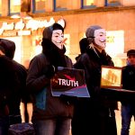 A group of people are gathered wearing Guy Fawkes masks holding signs that say “Truth.”