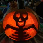 close up of an orange pumpkin carved into a skeleton-like figure with a goofy face. It is lit with a candle from the inside in a dark room sitting on a table. This is a close up of an orange pumpkin carved into a skeleton-like figure with a goofy face. It is lit with a candle from the inside in a dark room sitting on a table.