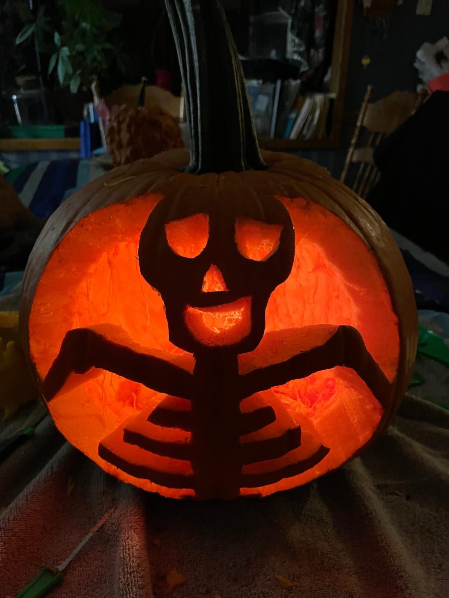 close up of an orange pumpkin carved into a skeleton-like figure with a goofy face. It is lit with a candle from the inside in a dark room sitting on a table. This is a close up of an orange pumpkin carved into a skeleton-like figure with a goofy face. It is lit with a candle from the inside in a dark room sitting on a table.
