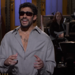 A man in a blue collar, gold chain, and sunglasses speaking in front of a band