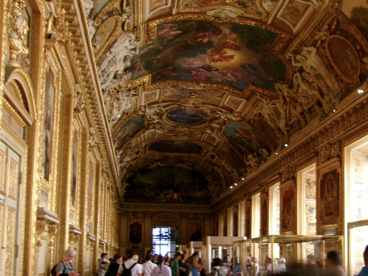 A crowd of people stand inside of the Galerie d'Apollon. Several paintings are on its ceiling and walls.