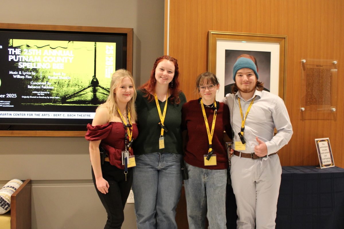 Four people with yellow lanyards pose in front of a theater display for The 25th Annual Putnam County Spelling Bee.