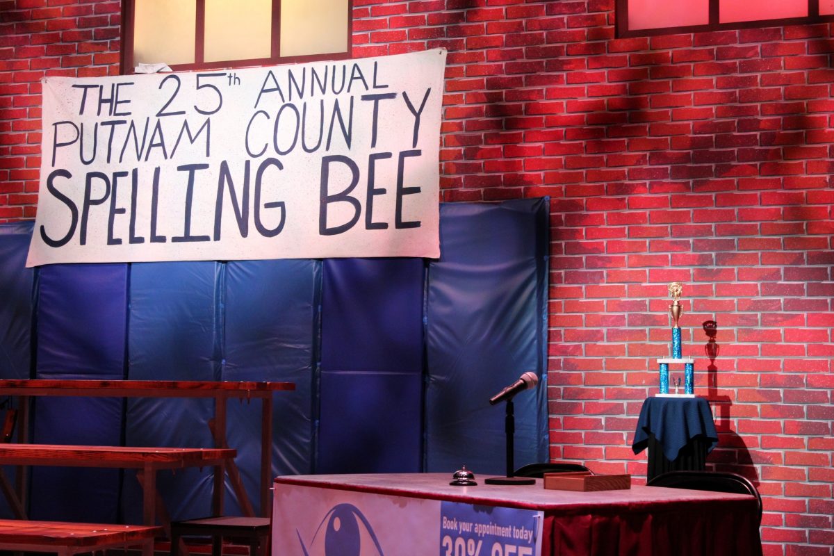 A sign saying "The 25th Annual Putnam County Spelling Bee" hanging from a stage decorated like a school gym.