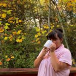 A person wearing a pink button-up shirt stands on a wooden deck surrounded by autumn trees with yellow and orange leaves. They’re sipping from a white mug.