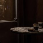 Two cups of coffee sit on a table with dim lights from outside coming through the window.