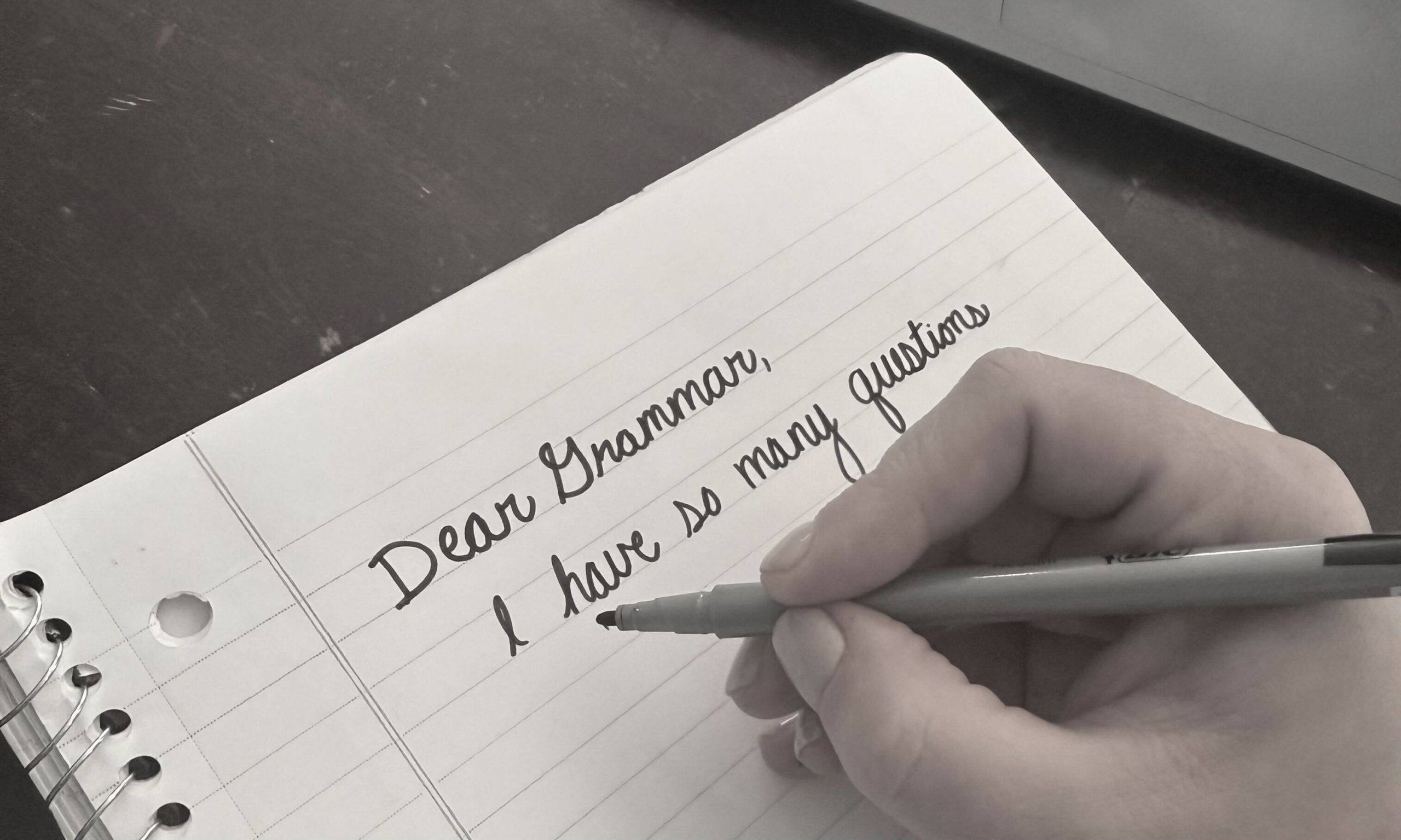 A hand holding a marker near notebook paper. The paper reads Dear Grammar, I have so many questions."