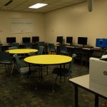 A room with several computers and a printer. A few chairs surround the two tables in the middle of the room.