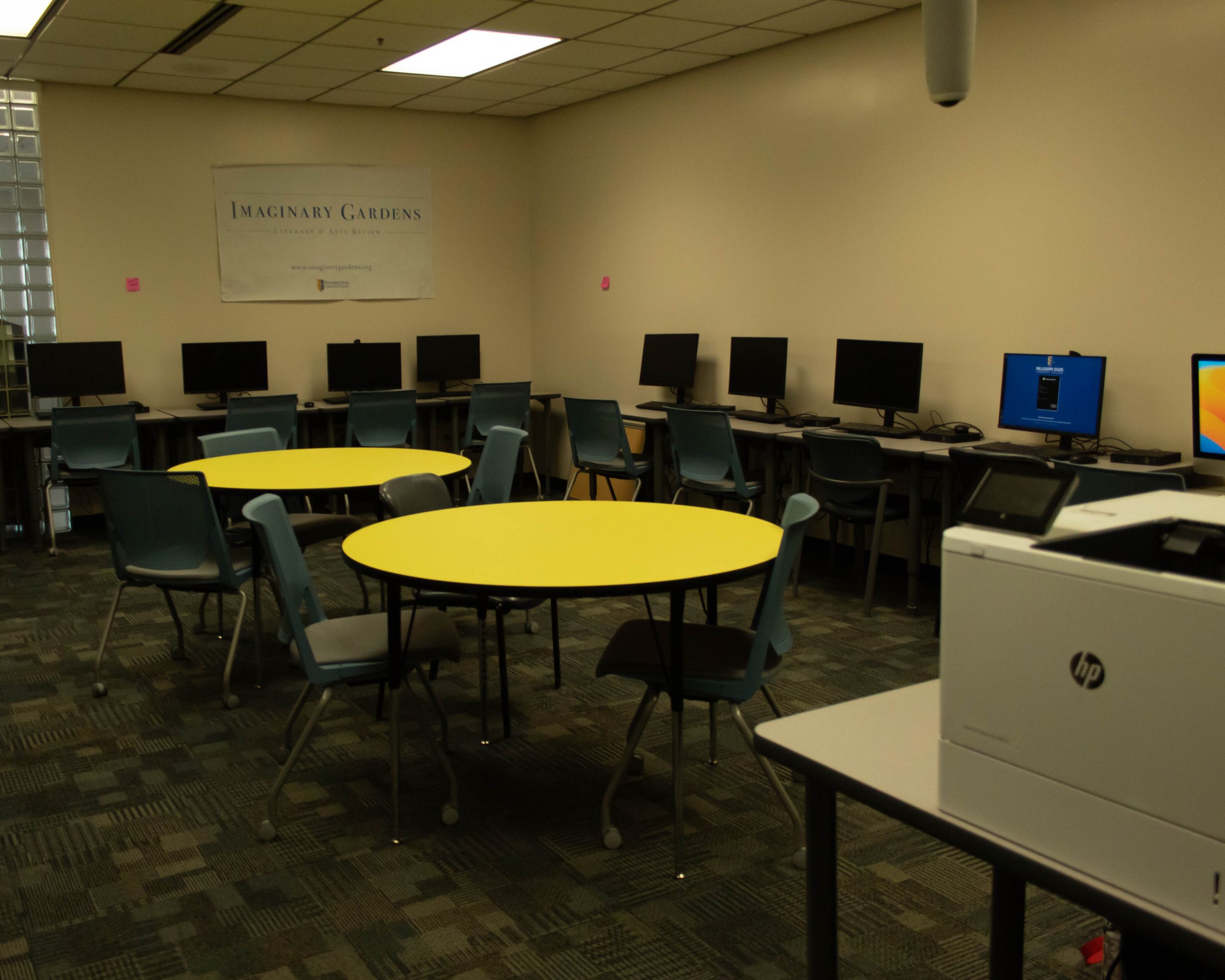 A room with several computers and a printer. A few chairs surround the two tables in the middle of the room.
