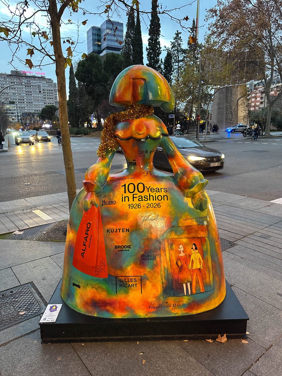 A rainbow sculpture of a doll-like woman with multiple fashion brands written as well as “100 years in Fashion 1926-2026” on the front of her dress