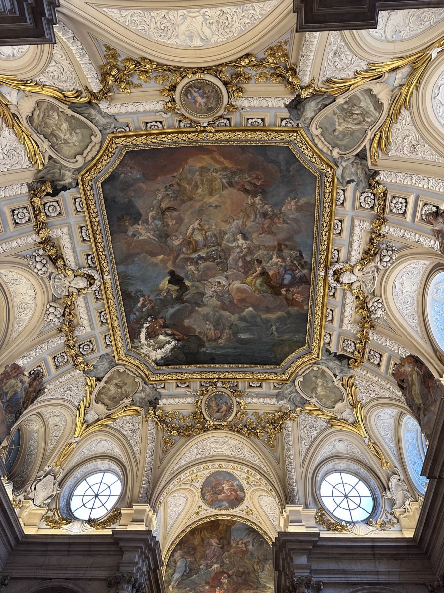 A white ceiling embellished with gold accents, windows, and a classical style mural in the center