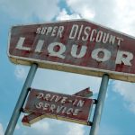 Against the clouds stands a red sign advertising for drive-in service at "Super Discount Liquor"