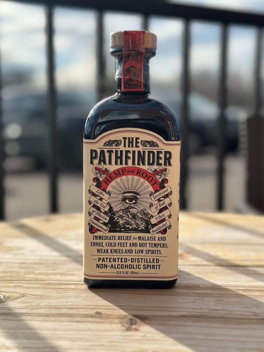 On a wooden table stands a non-alcoholic liquor bottle named The Pathfinder which is labeled with a black and white eye above a mountain peak