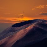 Birds fly over a crater. The sky is partly cloudy and orange.