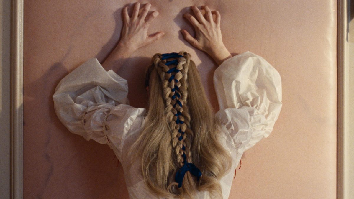 A person with long blond hair braided with a blue ribbon, wearing a white blouse, presses their hands against a pink padded surface.