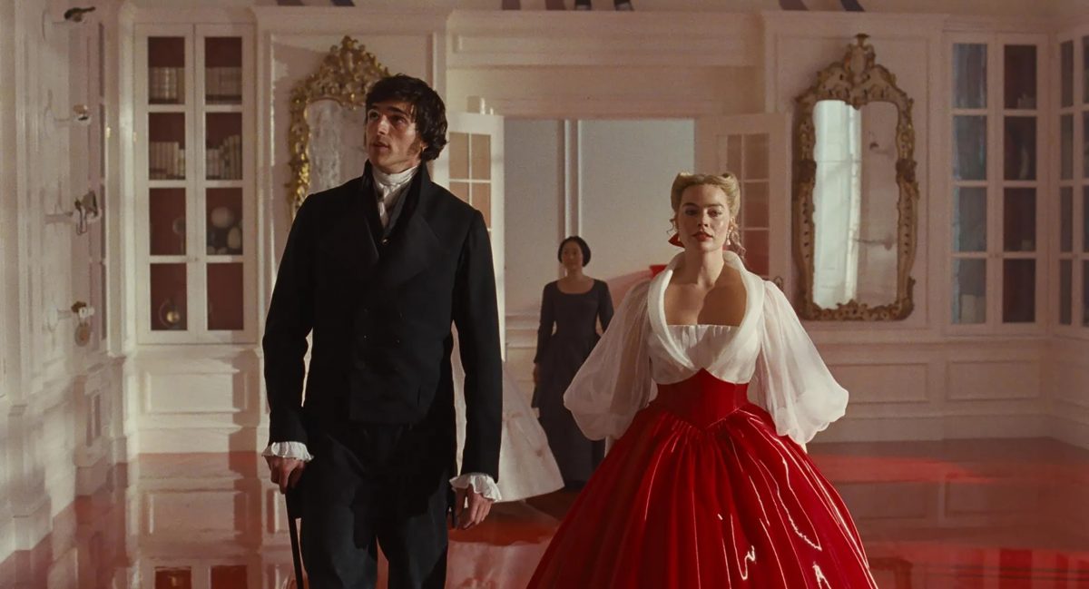 A man in a black suit and a woman in a red and white gown walk through an ornate room with mirrors and gold accents, while another woman stands in the background.