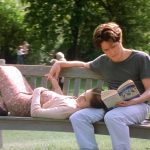 Anna and William hold hands on a bench at the park. Anna is lying down, while William is sitting and reading a book.