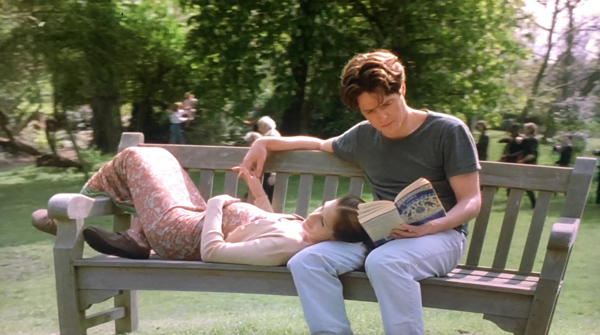 Anna and William hold hands on a bench at the park. Anna is lying down, while William is sitting and reading a book.