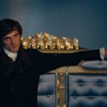 A man in 19th-century formal attire reclines with one arm draped over the back of an ornate blue and gold sofa, gazing forward in a softly lit, elegant room.