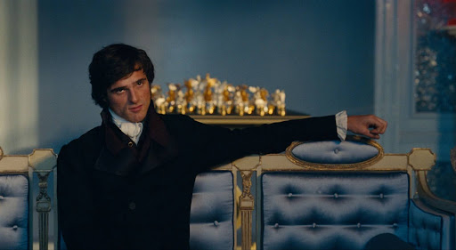 A man in 19th-century formal attire reclines with one arm draped over the back of an ornate blue and gold sofa, gazing forward in a softly lit, elegant room.
