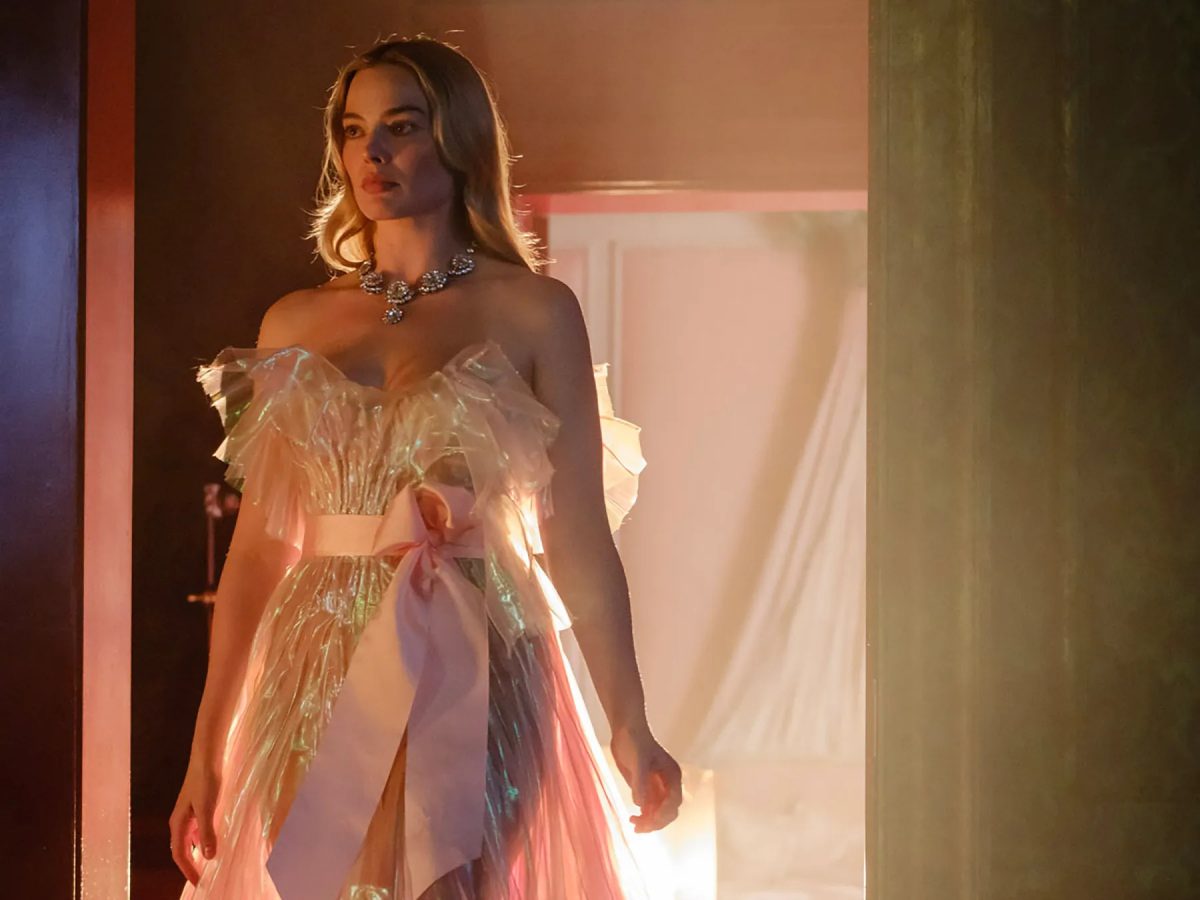 A woman in a shimmering, off-the-shoulder gown with a large pink bow at the waist steps through a doorway, wearing a statement necklace and lit by warm, golden light.
