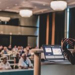 Person standing in front of a room, giving a presentation to a crowd
