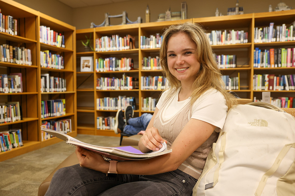 A college-aged girl sitting in a library, smiling at the camera as she flips through a notebook.