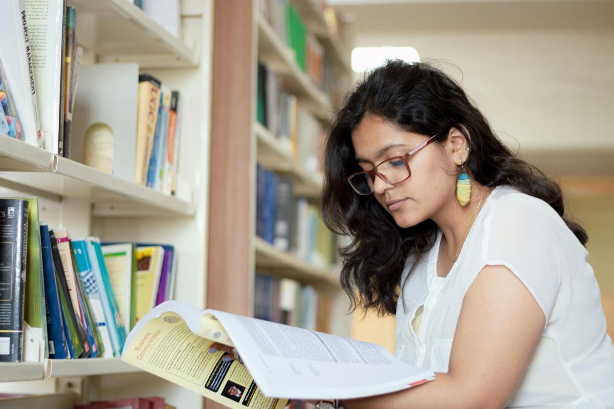 A young woman reading a book with bookshelves behind her.