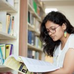 A young woman reading a book with bookshelves behind her.