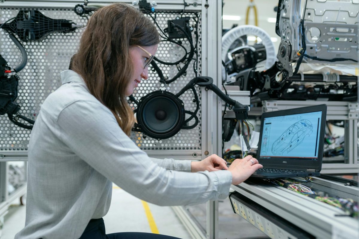 A woman on a laptop with schematics pulled up, in a workshop surrounded by machines.